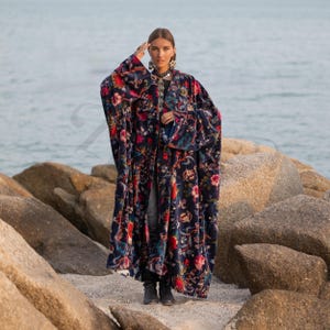 May include: A long, flowing robe with a dark blue background and a vibrant floral pattern in red, pink, and green. The robe has wide sleeves and is worn open. The garment is made of a soft, velvety material. The robe is a statement piece.