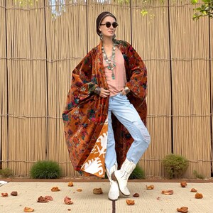 Model wearing the Amber Flora Harper Jade Velvet Kimono Robe by ZinniaPearl, standing on a woven mat with rustic bamboo fencing in the background. The plus size boho duster features burnt sienna cotton velvet with magenta, teal, and emerald florals, oversized sleeves, and deep pockets. Styled with layered necklaces, distressed denim, and white boots, this handcrafted robe celebrates autumn tones, bohemian elegance, and inclusive sizing from M to 4X.