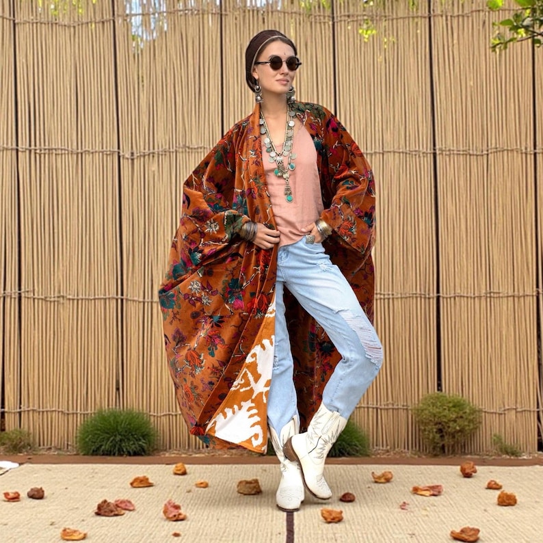 Model wearing the Amber Flora Harper Jade Velvet Kimono Robe by ZinniaPearl, standing on a woven mat with rustic bamboo fencing in the background. The plus size boho duster features burnt sienna cotton velvet with magenta, teal, and emerald florals, oversized sleeves, and deep pockets. Styled with layered necklaces, distressed denim, and white boots, this handcrafted robe celebrates autumn tones, bohemian elegance, and inclusive sizing from M to 4X.