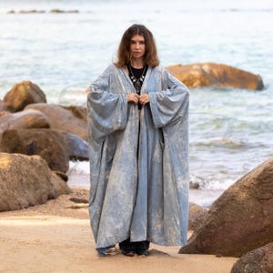 May include: A light blue, open-front robe with wide sleeves. The robe has a textured, almost tie-dye appearance. The person is standing on a sandy beach with rocks and the ocean in the background. The robe is a type of outerwear.