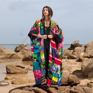 Puede incluir: Un kimono largo, colorido y de patchwork con una base negra y cierre de nudo. El kimono tiene un patrón de teñido anudado con tonos de azul, verde, rosa y amarillo. La modelo lleva el kimono sobre una blusa negra y pantalones vaqueros.
