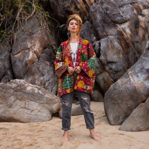 May include: A long-sleeved, open-front jacket featuring a vibrant patchwork of red, yellow, and green floral and geometric patterns. The garment is worn over a plain white top and distressed charcoal gray denim jeans with ripped knees.