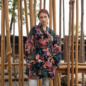 Close-up portrait of the model in the Mei Velvet Kimono Duster – Nocturne Flora. Set among tall bamboo poles, the image showcases the velvet’s lush floral design and the model’s confident, soulful expression. Her layered jewelry and relaxed styling enhance the jacket’s boho-chic mood, emphasizing ZinniaPearl’s essence — artistry, inclusivity, and timeless, handcrafted elegance.