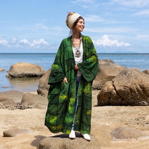 May include: A long, open-front green kimono featuring a dark grid pattern and yellow tie-dye accents. The garment has wide sleeves, two front pockets, and is worn over a white top and blue jeans, paired with a white head wrap and silver necklace.