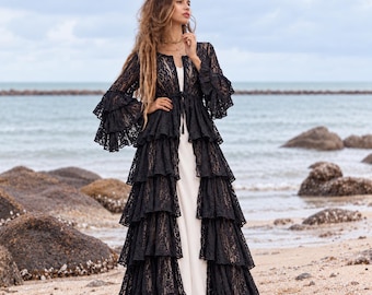May include: A floor-length black lace duster featuring long tiered ruffled sleeves and a tiered ruffled skirt, worn over a simple white sleeveless dress. The black lace garment has a front tie closure at the waist and a round neckline.