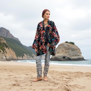 Model standing barefoot on a sandy beach wearing the Mei Velvet Kimono Duster in Nocturne Flora by ZinniaPearl. The black velvet jacket features rich floral embroidery in rust, magenta, jade, and amber tones, styled with patterned denim and boho jewelry. A rocky coastline and sea cliffs create a dreamy, nature-inspired backdrop that highlights the artisanal luxury and free-spirited bohemian elegance of the design.