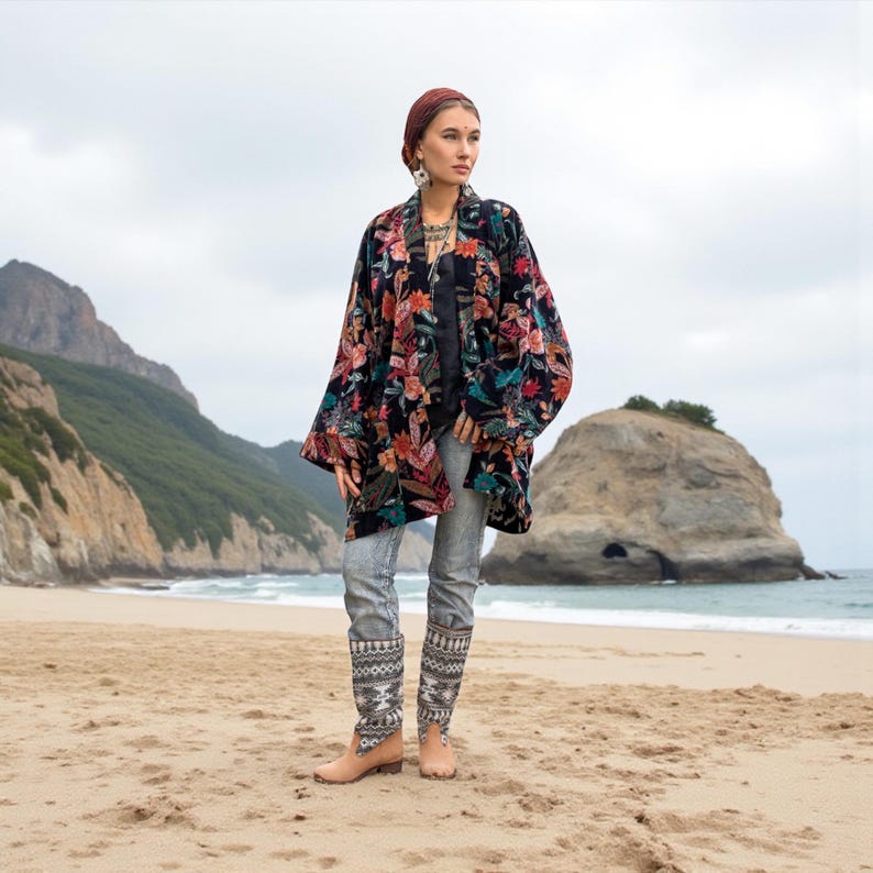 Model standing barefoot on a sandy beach wearing the Mei Velvet Kimono Duster in Nocturne Flora by ZinniaPearl. The black velvet jacket features rich floral embroidery in rust, magenta, jade, and amber tones, styled with patterned denim and boho jewelry. A rocky coastline and sea cliffs create a dreamy, nature-inspired backdrop that highlights the artisanal luxury and free-spirited bohemian elegance of the design.