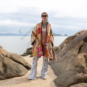 以下が含まれることがあります： ベージュ、オレンジ、ピンク、赤の色合いのパッチワークの着物風ジャケット。 ジャケットはゆったりとしたフィット感と長袖です。 ライトブルーのダメージジーンズとサングラスを着用しています。 背景にはビーチと海があります。