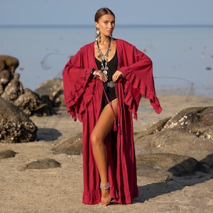 May include: A long, flowing red kimono-style robe with ruffled sleeves and a tiered skirt. The robe is open in the front, revealing a black swimsuit and layered necklaces. The model is standing on a sandy beach with rocks and the ocean in the background.