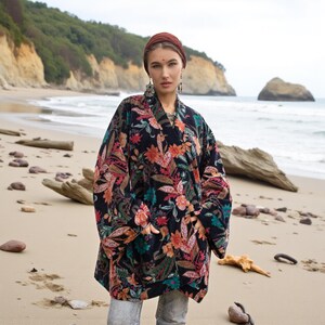 Close-up portrait of the Mei Velvet Kimono Duster in Nocturne Flora by ZinniaPearl. The model stands on a tranquil beach wearing bold statement earrings and a headscarf, her hands tucked into the deep velvet pockets. The floral pattern glows with magenta, copper, and green embroidery against a black velvet backdrop, expressing timeless boho luxury and handcrafted beauty.