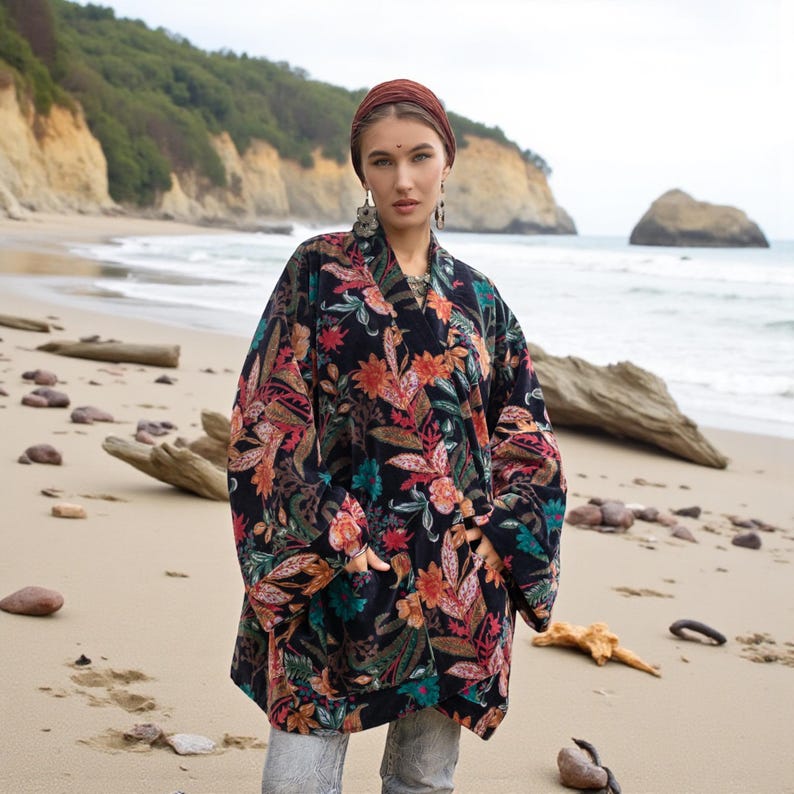 Close-up portrait of the Mei Velvet Kimono Duster in Nocturne Flora by ZinniaPearl. The model stands on a tranquil beach wearing bold statement earrings and a headscarf, her hands tucked into the deep velvet pockets. The floral pattern glows with magenta, copper, and green embroidery against a black velvet backdrop, expressing timeless boho luxury and handcrafted beauty.