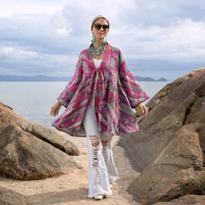 May include: A colourful patterned jacket in pink, green, and purple, with a tie closure. The jacket is worn with distressed white jeans. The outfit is accessorized with a necklace and sunglasses. The background shows a beach and the sea.