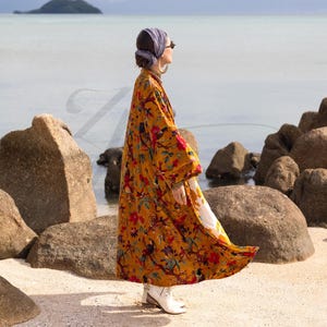 Side profile of the Golden Sunbird velvet duster showing the garment in motion. The rich cotton velvet fabric drapes elegantly, revealing the side seam and the volume of the wide sleeves. The golden mustard color contrasts with the blue ocean background. A faint "Z" logo watermark overlays the image. The model looks out toward the water, highlighting the sophisticated resort-wear aesthetic of this long bohemian coat.