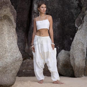 Könnte beinhalten: Ein zweiteiliges Outfit in Weiß, bestehend aus einem Crop-Top und einer Haremshose. Das Crop-Top hat dünne Träger. Die Haremshose ist locker geschnitten mit einem Patchwork-Design und Spitzendetails. Das Outfit wird an einem Sandstrand getragen.