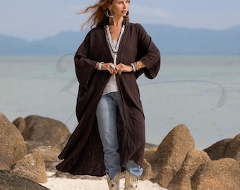 May include: A long, dark brown kimono-style robe with wide sleeves. The robe has a textured appearance and is worn over a white top and blue jeans. The model is wearing white cowboy boots with a star design. The background features a beach and ocean.