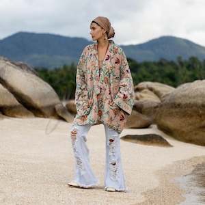 May include: A light pink floral print kimono with long sleeves and pockets. The kimono is paired with light wash, distressed flare jeans. The outfit is worn on a sandy beach with a mountain backdrop.