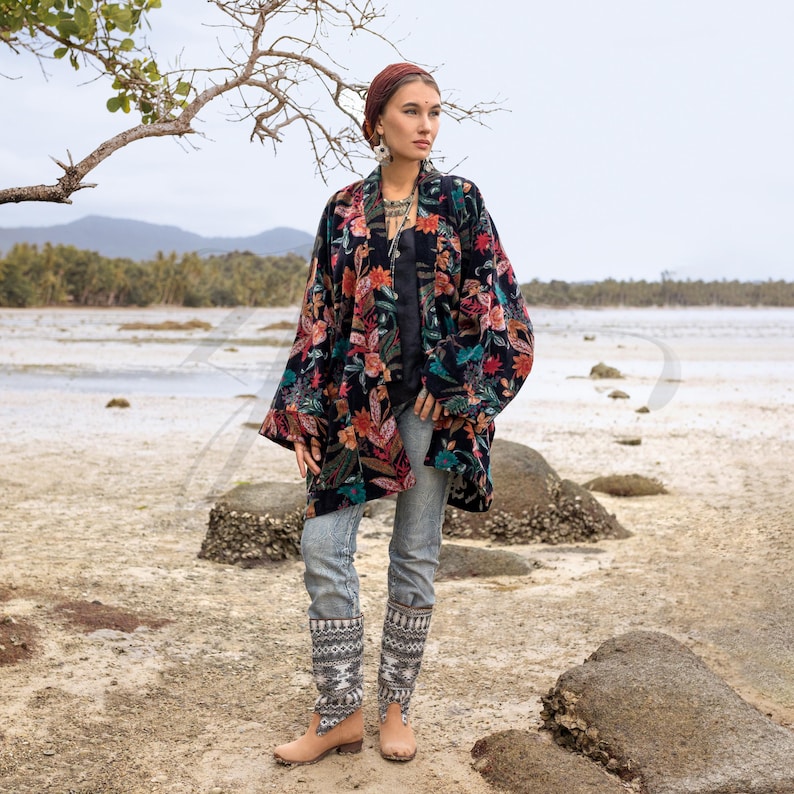 Model wearing the Mei Velvet Kimono Duster in Nocturne Flora by ZinniaPearl, standing on a rocky coastal shore with soft sea light. The black cotton-velvet jacket blooms with intricate florals in copper, magenta, jade green and amber. Styled with artisan denim and boho jewelry, this image captures the spirit of ZinniaPearl — free-spirited, handcrafted, and radiating timeless bohemian elegance.