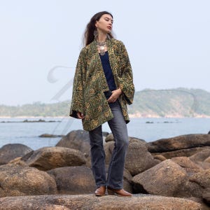Model standing on seaside rocks wearing an emerald green and gold hand block-printed velvet kimono jacket with wide sleeves and relaxed fit. Styled with jeans, brown boots, and silver jewelry, the boho jacket captures ZinniaPearl’s artisan elegance against a coastal backdrop.