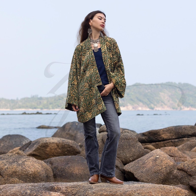 Model standing on seaside rocks wearing an emerald green and gold hand block-printed velvet kimono jacket with wide sleeves and relaxed fit. Styled with jeans, brown boots, and silver jewelry, the boho jacket captures ZinniaPearl’s artisan elegance against a coastal backdrop.