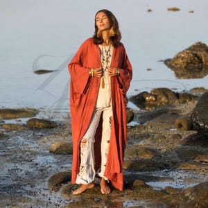 Könnte beinhalten: Ein langer, fließender Kimono-Stil-Robe in Rostrot mit weiten Ärmeln. Die Robe ist vorne offen und zeigt ein weißes Oberteil und zerrissene weiße Jeans. Die Person trägt mehrere Halsketten und Ohrringe und steht an einem felsigen Strand.