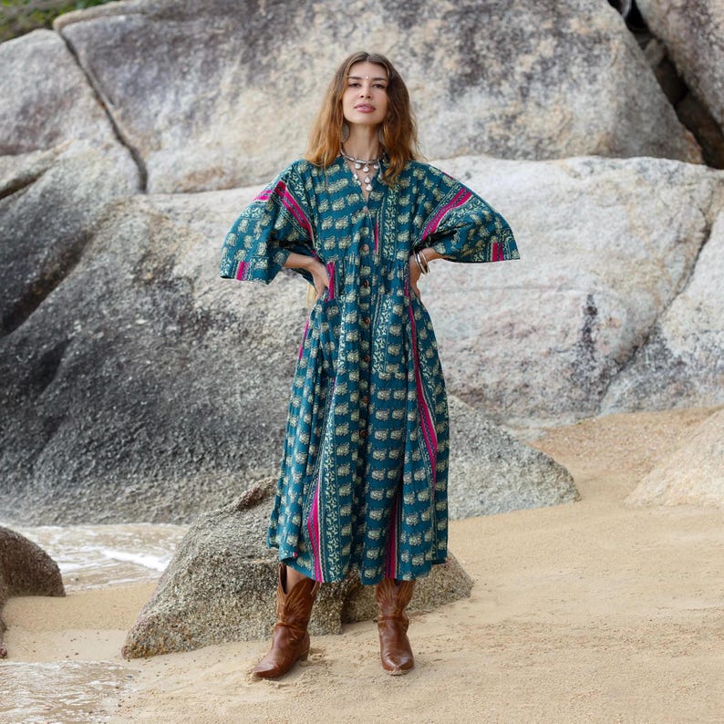 Woman modeling the Cora Midi Dress in Peacock Rose on a rocky beach landscape. The oversized boho midi dress features teal and magenta artisan print details, flowing wide kimono sleeves, and a relaxed babydoll silhouette styled with brown leather boots. The lightweight cotton fabric drapes beautifully with effortless movement, creating a free-spirited bohemian look perfect for coastal styling, festivals, and everyday artisan fashion.