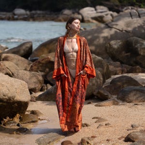 May include: A long, flowing orange and brown kimono with a floral pattern. The kimono is worn on a beach with rocks and water in the background. The garment has wide sleeves and a loose fit, perfect for a beach cover-up or resort wear.