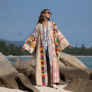 Könnte beinhalten: Ein langer, beigefarbener Kimono-Stil-Mantel mit farbenfrohen Patchwork- und Stickdetails. Der Mantel weist eine Vielzahl geometrischer Muster und lebendiger Farben auf, darunter Rot, Gelb und Blau. Die Ärmel und Kanten sind mit einem zarten, strukturierten Stoff verziert.