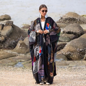 May include: A long, open-front kimono-style robe featuring a patchwork design of various black, white, and multi-colored patterned fabrics. The garment has wide sleeves and a loose, flowing silhouette, worn over a simple white top and light-colored shoes.