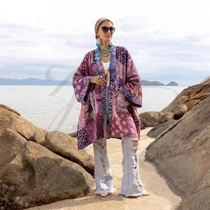 May include: A patchwork kimono in shades of purple, pink, and blue. The kimono features a variety of floral and geometric patterns. The garment has long sleeves and pockets. The person is wearing light blue ripped jeans.