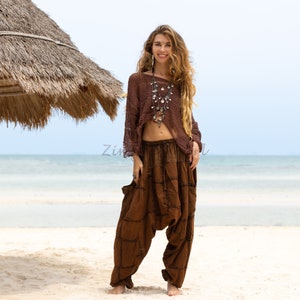 May include: A woman wearing a brown, open-weave knit top and matching brown harem pants with large pockets. The pants have a patchwork design. She is wearing a long silver necklace and standing on a sandy beach with a thatched roof in the background.