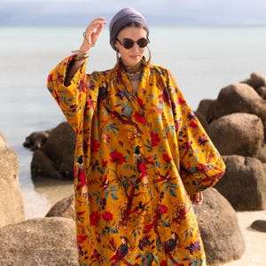 Bohemian style mustard yellow velvet kimono duster worn by a model on a rocky beach. The "Golden Sunbird" print features vibrant red flowers and colorful birds on a gold background. The model wears a purple headwrap, round sunglasses, and hoop earrings, holding one hand near her head. The loose-fitting cotton velvet robe features wide sleeves and an open front, showcasing the luxurious drape and rich fabric texture against a calm ocean backdrop.