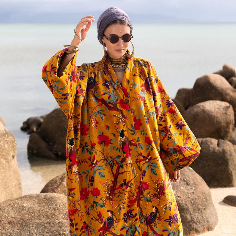 Bohemian style mustard yellow velvet kimono duster worn by a model on a rocky beach. The "Golden Sunbird" print features vibrant red flowers and colorful birds on a gold background. The model wears a purple headwrap, round sunglasses, and hoop earrings, holding one hand near her head. The loose-fitting cotton velvet robe features wide sleeves and an open front, showcasing the luxurious drape and rich fabric texture against a calm ocean backdrop.