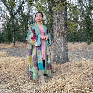 May include: A woman wearing a long, patchwork quilt jacket in shades of blue, green, brown, and red. The jacket has a quilted texture and a loose fit. She is standing in a field of dry grass.
