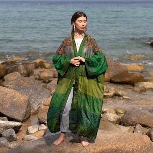 May include: A long, flowing green and brown kimono-style robe with a floral pattern. The robe has wide sleeves and is worn open. The model is standing on a rocky beach near the ocean.