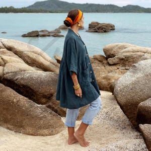 Side profile of the Smoky Jade Aria Tunic showing the soft drape of breathable crinkle cotton gauze fabric. The oversized boho silhouette with dropped shoulders and wide sleeves creates graceful movement as the model walks barefoot along a rocky beach path. Paired with rolled denim jeans, the tunic demonstrates relaxed resort styling and airy comfort.