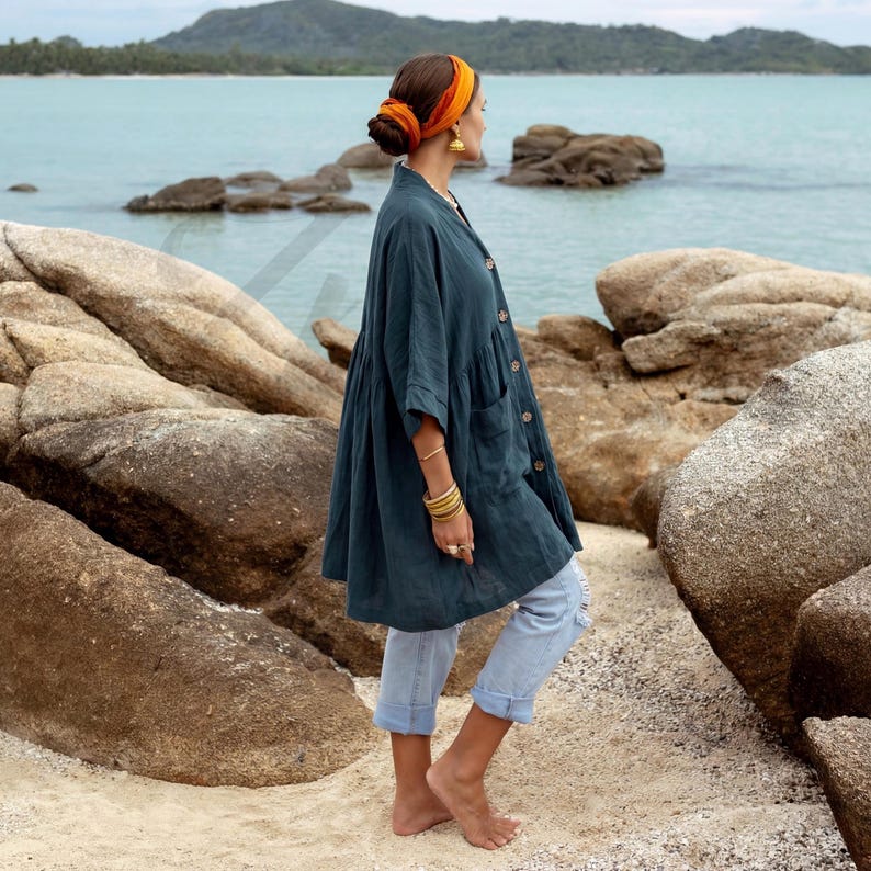 Side profile of the Smoky Jade Aria Tunic showing the soft drape of breathable crinkle cotton gauze fabric. The oversized boho silhouette with dropped shoulders and wide sleeves creates graceful movement as the model walks barefoot along a rocky beach path. Paired with rolled denim jeans, the tunic demonstrates relaxed resort styling and airy comfort.