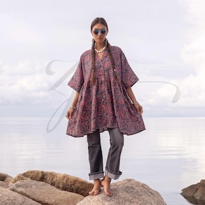 Op de afbeelding: Een ruimvallende tuniek met knoopsluiting, voorzien van een donkerrood en blauw paisley-patroon, korte wijde mouwen en een uitlopend silhouet, gecombineerd met een rechte grijze spijkerbroek met omgeslagen zomen voor een casual uitstraling.
