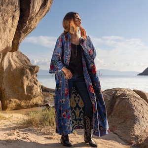 May include: A long blue floral patterned robe with a black top and blue jeans. The person is standing on a sandy beach with large rocks in the background.