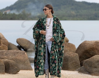 May include: A long, dark green velvet kimono with a floral pattern in shades of red, pink, and yellow. The kimono is worn over a white top and blue jeans. The person is wearing white boots and a long necklace.