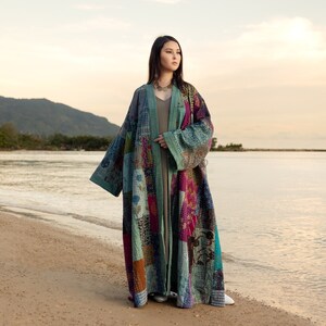 Puede incluir: Una mujer lleva un abrigo largo de estilo patchwork con una variedad de colores y patrones, incluyendo azul, verde, rosa y morado. Está de pie en una playa de arena con el océano de fondo.