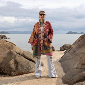 May include: A patchwork kimono-style jacket in shades of red, orange, brown, and green. The jacket has a unique design with pockets and a mix of patterns. The person is wearing ripped light wash jeans and accessories.