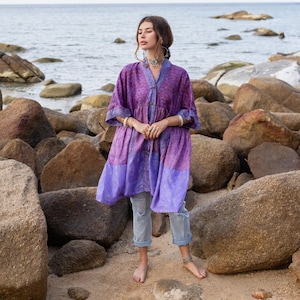 Peut inclure: Une tunique boutonnée violette et lavande avec un motif floral. La tunique a des manches trois-quarts et un ourlet à volants. Le modèle porte un jean bleu clair et des bijoux argentés. L'arrière-plan est une plage avec des rochers et l'océan.