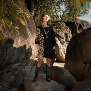 May include: A woman wearing a black dress with a tie-dye pattern on the right side, paired with black cowboy boots. She is also wearing a silver necklace with multiple pendants. The background features large rocks and foliage.