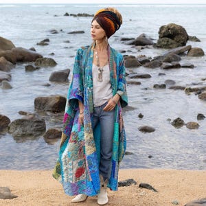 Peut inclure: Une femme portant un long kimono patchwork bleu et multicolore avec un débardeur blanc et un jean bleu. Elle se tient sur une plage avec des rochers et de l'eau en arrière-plan. Elle porte un turban coloré sur la tête.