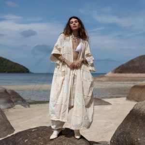 Peut inclure: Une femme portant une robe blanche longue et ample en coton avec un motif patchwork. Elle est debout sur un rocher avec la mer en arrière-plan.