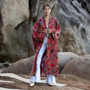 May include: A long, flowing, burgundy velvet kimono with a vibrant floral pattern in shades of red, green, and blue. The kimono is open in the front and worn over a white top and light blue jeans. The model is standing on a rocky surface.