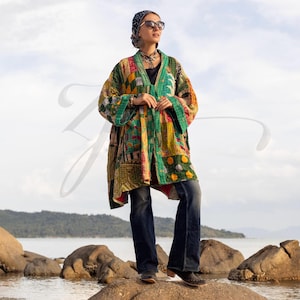 Op de afbeelding: Een patchwork kimono-achtige jas in groen, geel en rood. De jas heeft verschillende geometrische en bloemmotieven. De persoon draagt een donkerblauwe jeans, een zonnebril en een patroon hoofddoek.