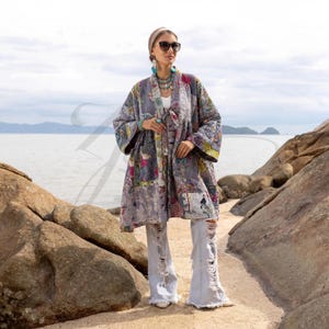 May include: A woman wearing a long, quilted gray kimono with colorful floral and patchwork designs. She is also wearing light-wash, ripped jeans, sunglasses, and turquoise jewelry. The background features a seascape with rocks and a cloudy sky.