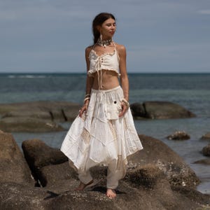 May include: A cream-colored two-piece outfit, featuring a halter top with a lace-up front and a flowing skirt with patchwork details. The outfit is worn with a matching choker and bracelets. The ensemble is perfect for a beach or bohemian style.