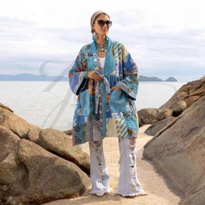 May include: A long, patchwork kimono in shades of blue, with floral and geometric patterns. The kimono has pockets and a tie closure. The person is wearing sunglasses, a white top, and distressed jeans. The background is a beach scene with rocks and water.